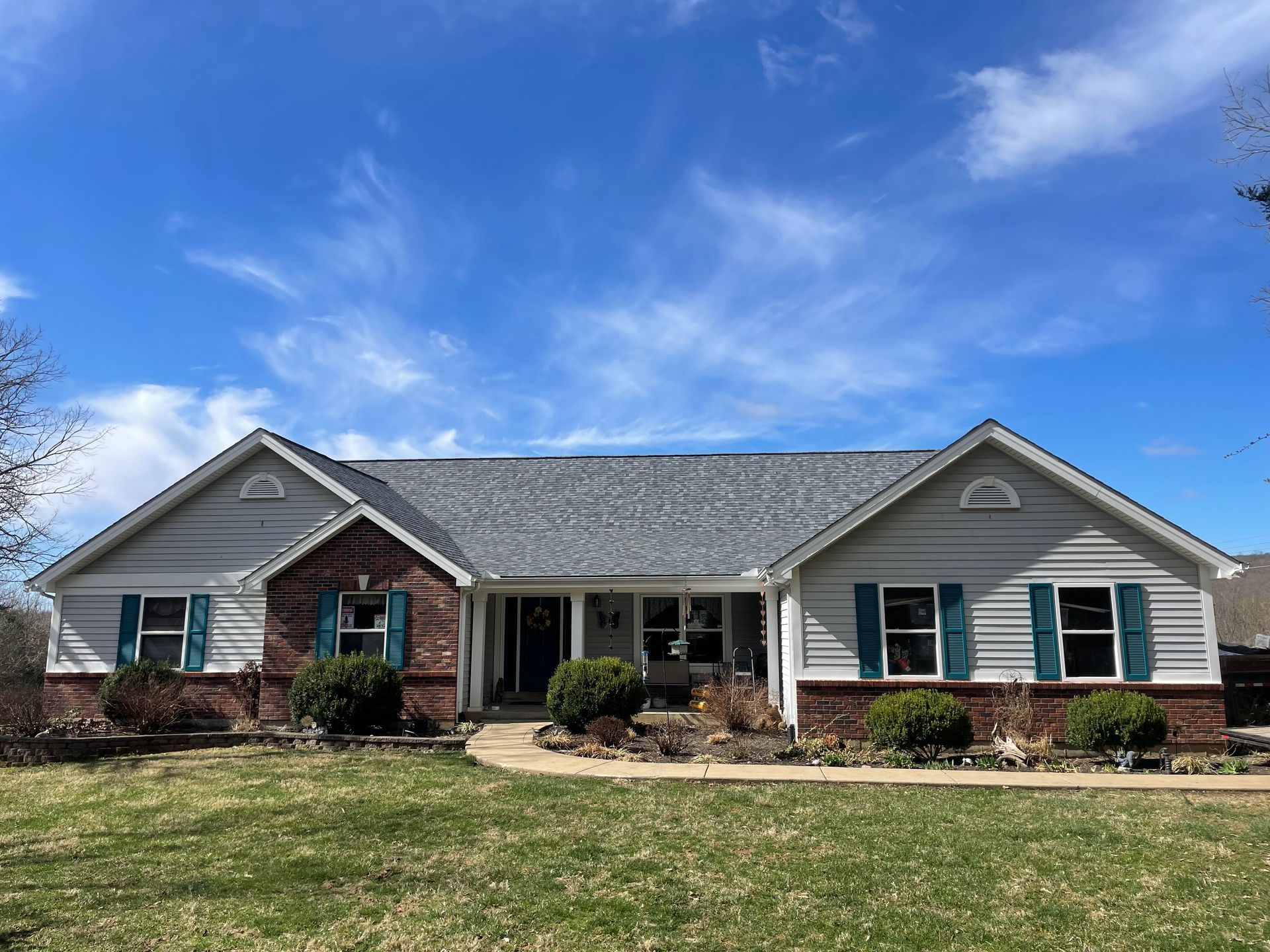 Ranch-style house with gray roof, light siding, brick accents, and turquoise shutters against a blue sky.