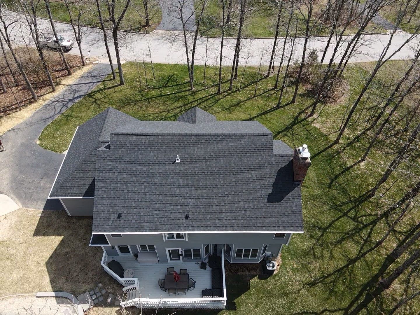 Aerial view of a gray house with a dark roof and a deck, surrounded by trees and a driveway.
