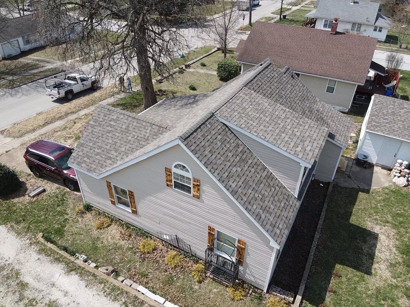 Aerial view of a gray house with brown shutters and a brown roof. A vehicle is parked in the driveway.