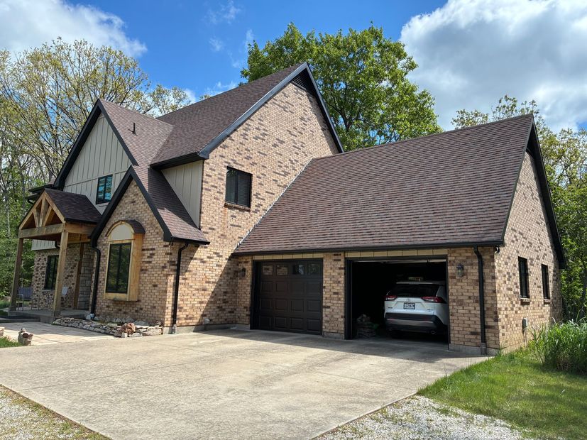 Two-story brick house with attached garage; dark brown roof, gray concrete driveway, blue sky. Car in garage.
