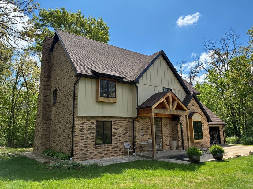 Two-story brick house with brown roof and light tan siding; porch with dark wood beams.