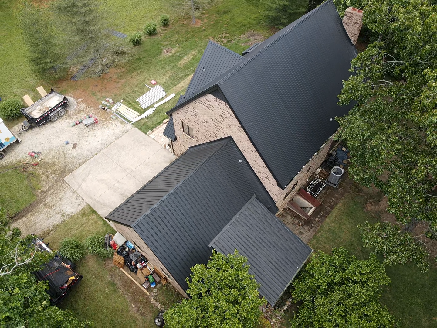 Aerial view of a house with a dark gray metal roof, brick exterior, and a concrete driveway surrounded by trees.