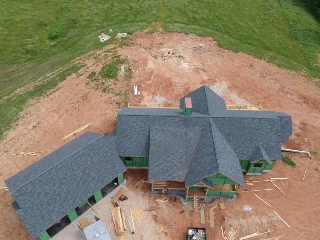 Aerial view of a house under construction with dark gray roof, green framing, surrounded by red dirt and grass.