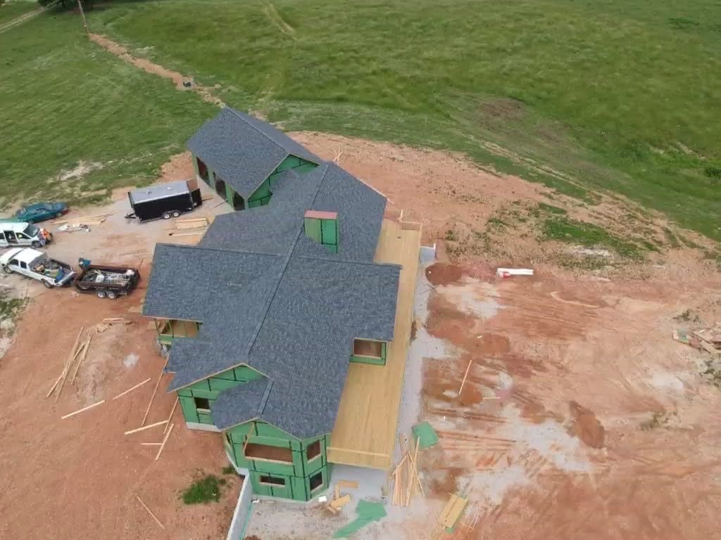 Aerial view of a house under construction with a dark gray roof, surrounded by dirt and green grass.