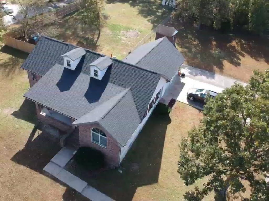 Aerial view of a two-story house with a gray roof and brick facade; a tree and a driveway are visible.
