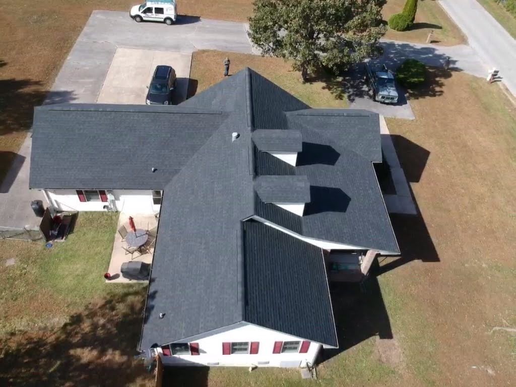 Aerial view of a house with a dark gray shingle roof, three dormers, and a driveway with vehicles.