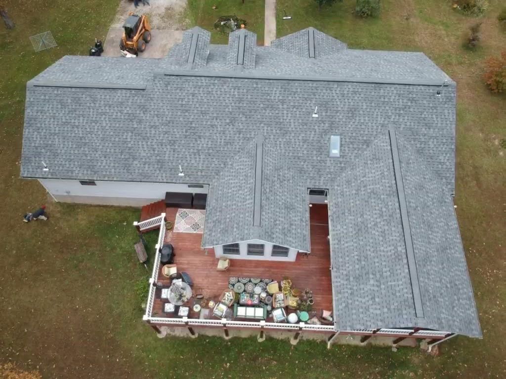 Aerial view of a house with a gray shingled roof, a large deck with furniture, and a grassy yard.