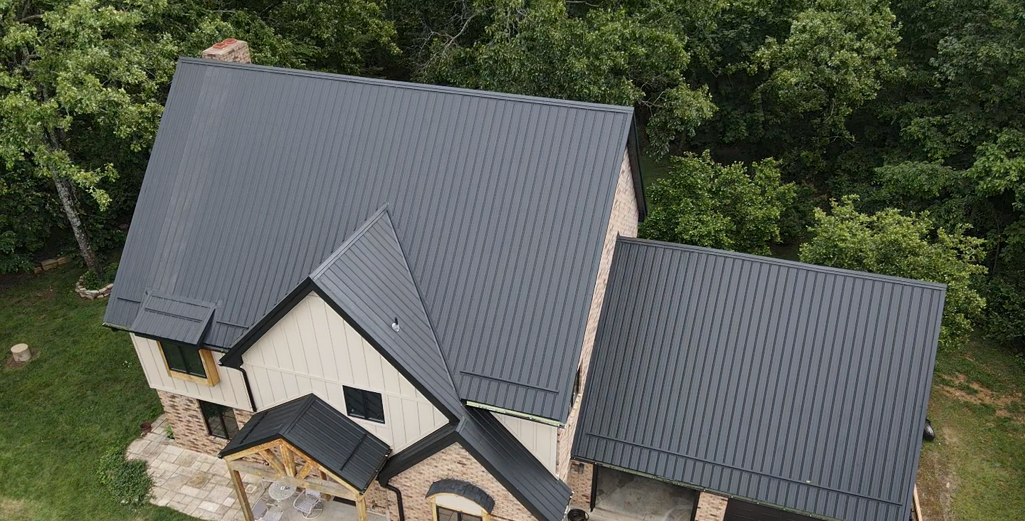 Dark gray metal roof on a two-story house with stone and beige siding, surrounded by trees.