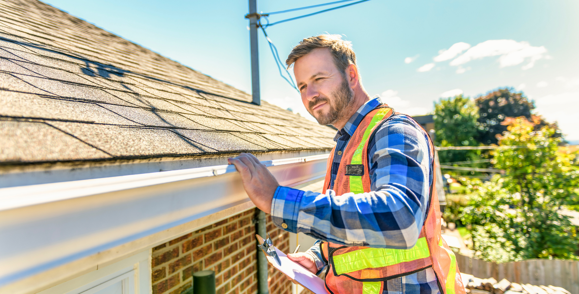 Man in safety vest inspecting a home's gutter and roof, holding a clipboard, sunny day.