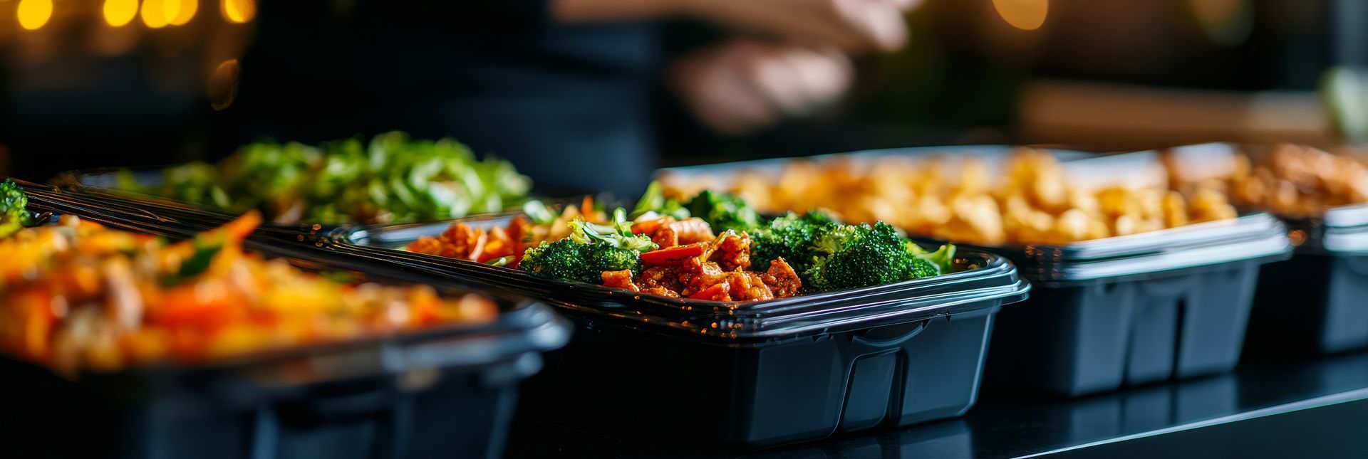 A low-angle view of multiple black takeaway containers filled with various colorful cooked meals on a dark counter.