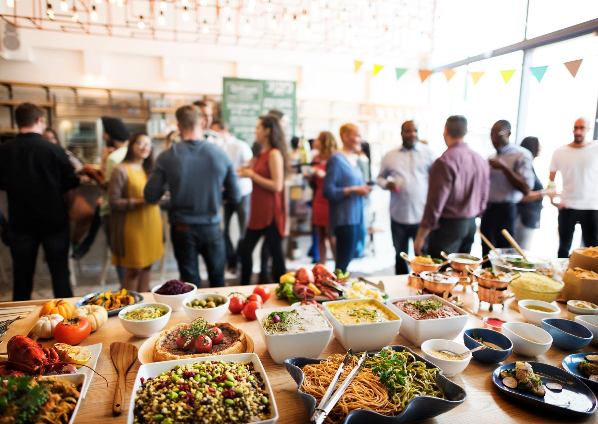 A spread of buffet food sits on a table in the foreground while a blurred group of people mingles in a bright, modern room.