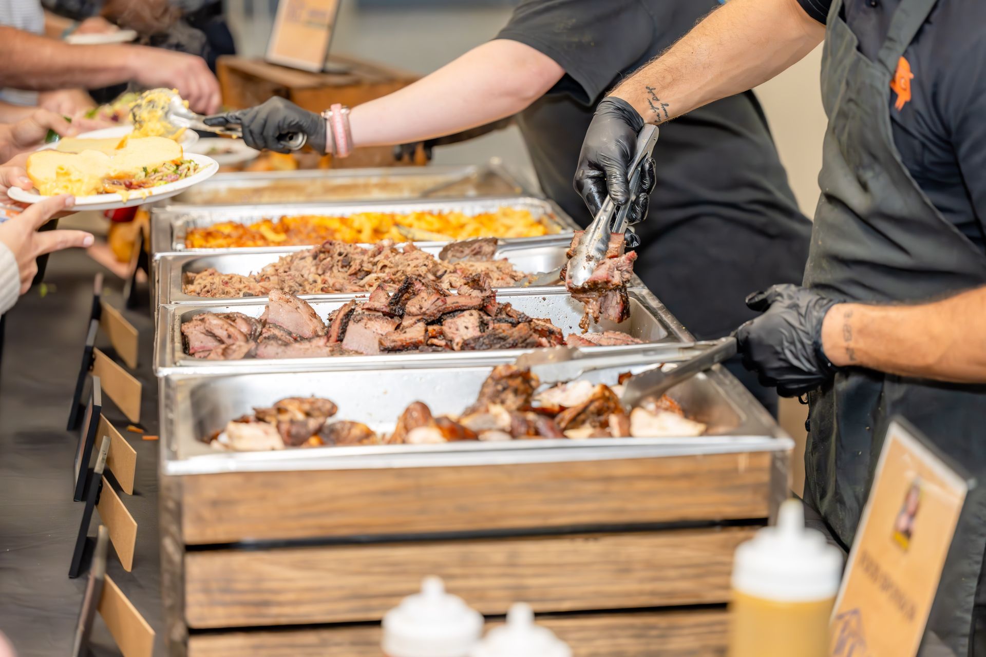 Workers in black uniforms and gloves serve meat and sides from a buffet line to guests at a catering event.
