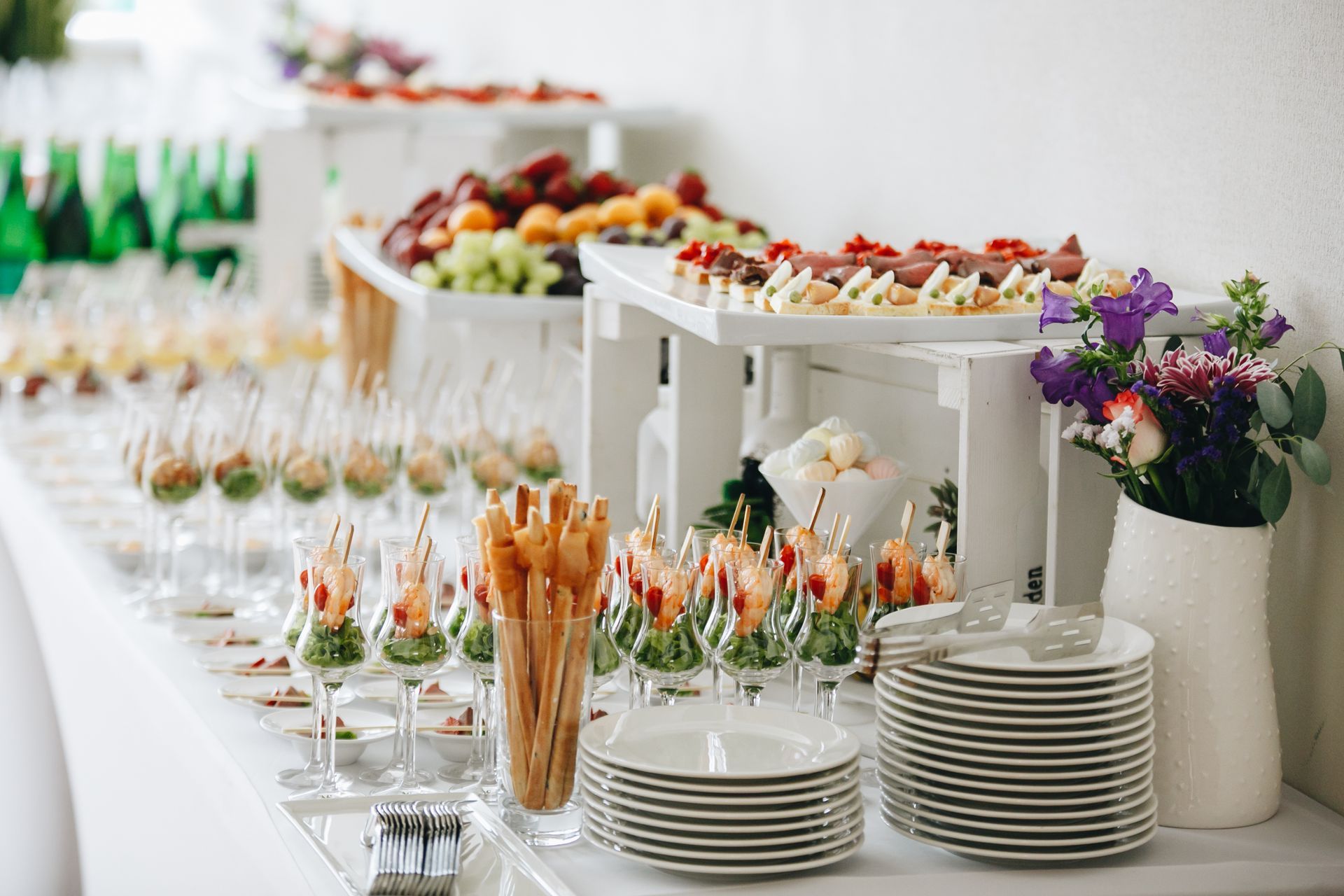 A buffet table set for an event with appetizer glasses, breadsticks, stacked plates, and a vase of flowers.