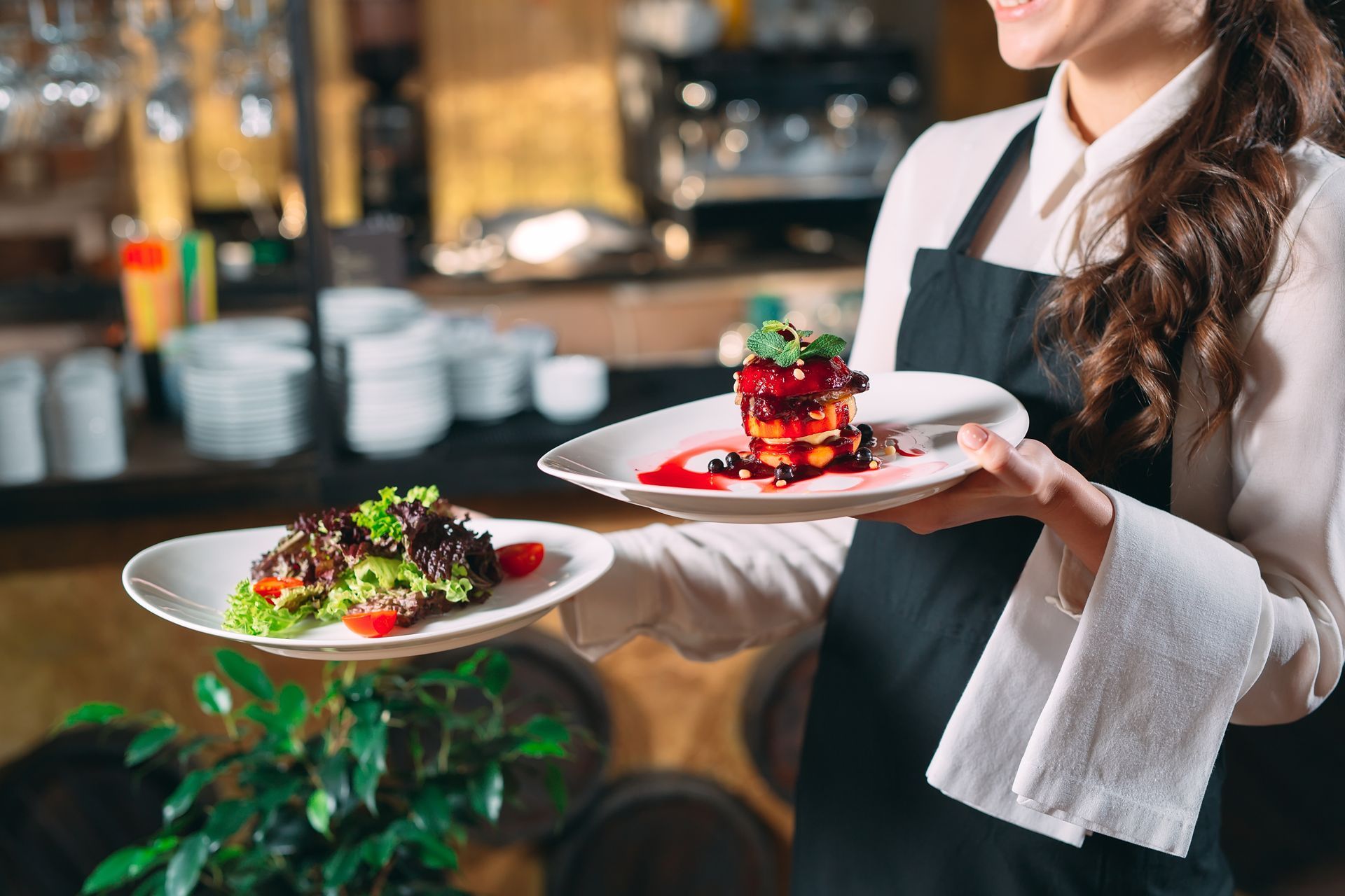 A server in a white shirt and black apron holds two plates of food in a restaurant setting.