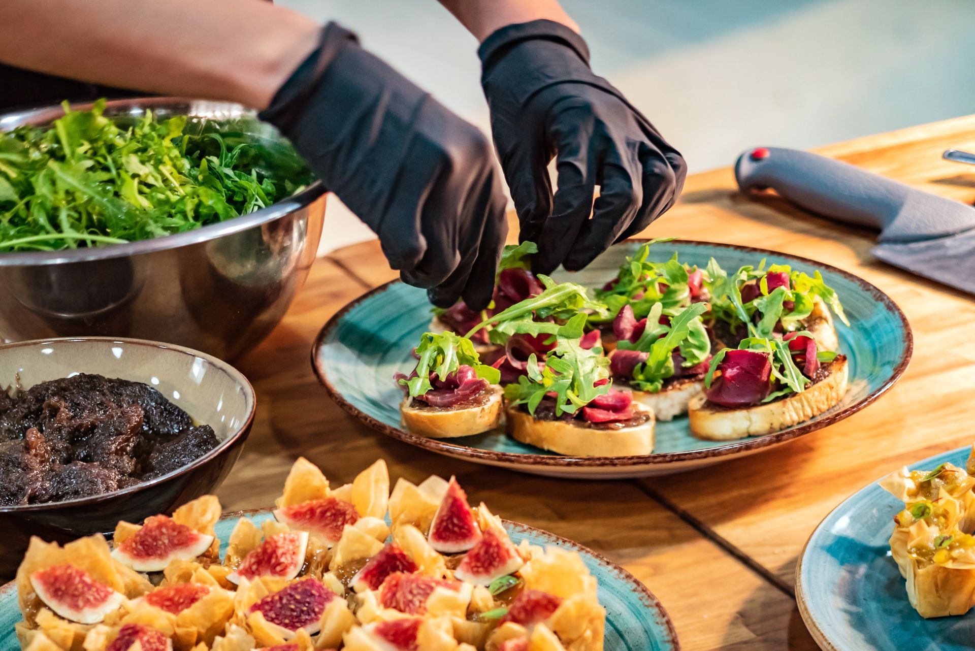 Hands wearing black gloves arrange arugula onto bruschetta on a plate, with bowls of ingredients on a wooden table.