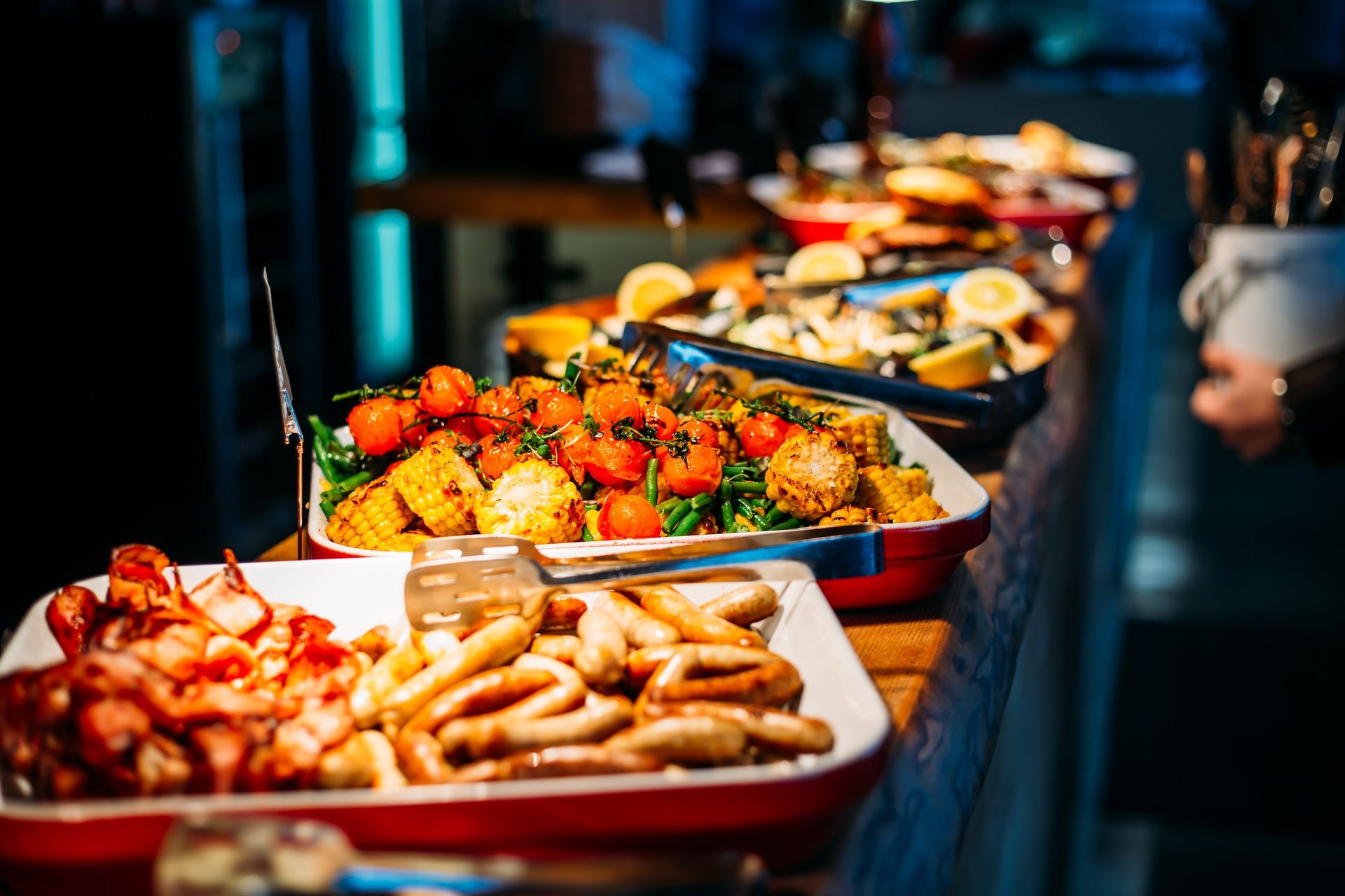 A buffet table featuring platters of cooked sausages, bacon, roasted tomatoes, and lemon-topped seafood in a dimly lit room.