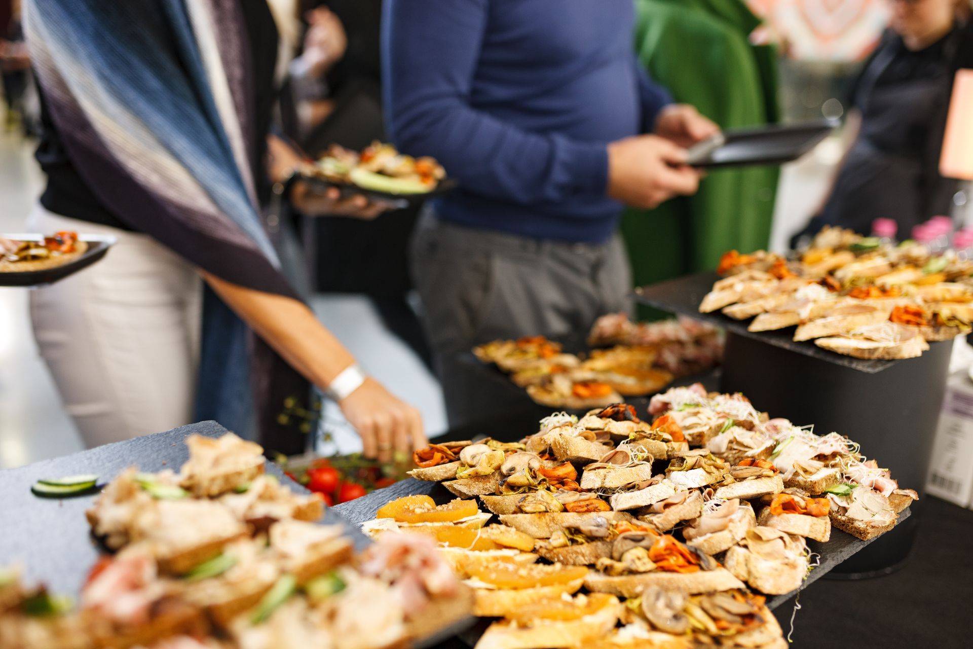 People at a catered event picking appetizers from platters of sandwiches, fruits, and snacks on a buffet table.