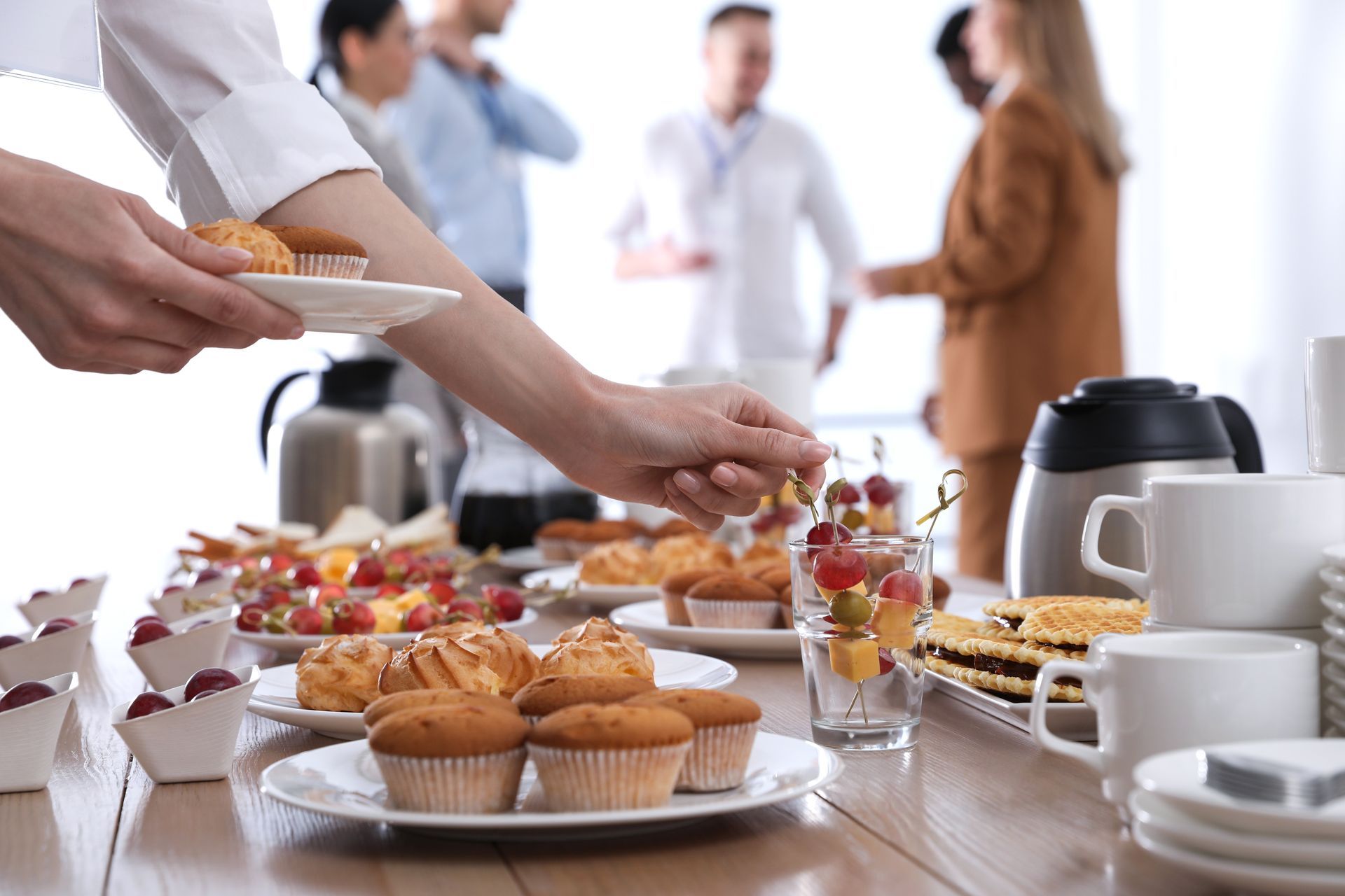 A person reaching for a snack at a coffee break catering table with a blurred group of people in the background.