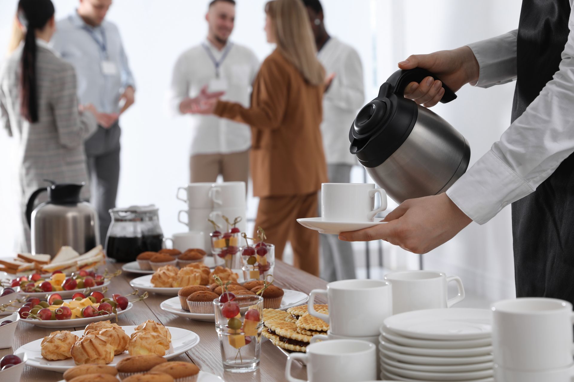 A person pours coffee into a cup at a catering table filled with snacks, with people socializing in the blurred background.