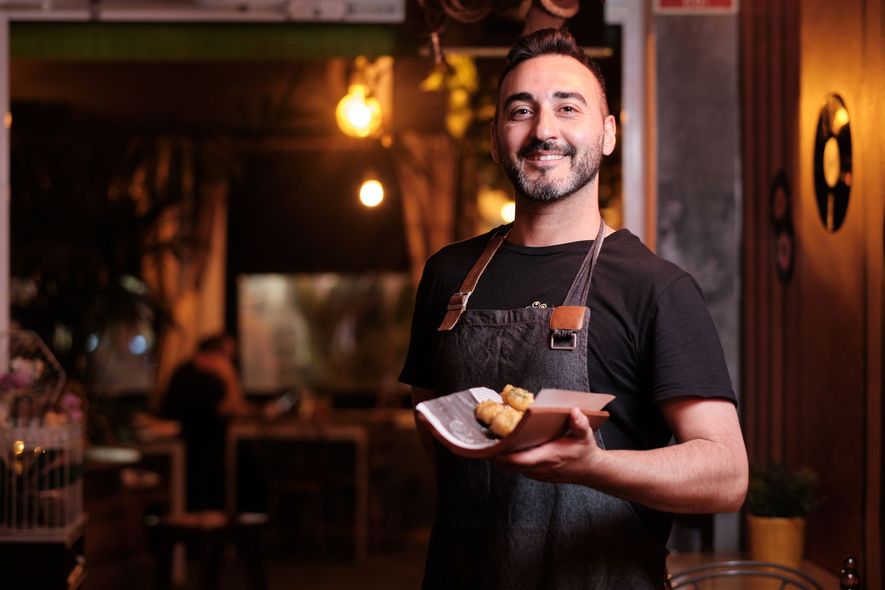 A smiling person in a black shirt and apron holds a small plate of food in a warmly lit, dimly restaurant setting.
