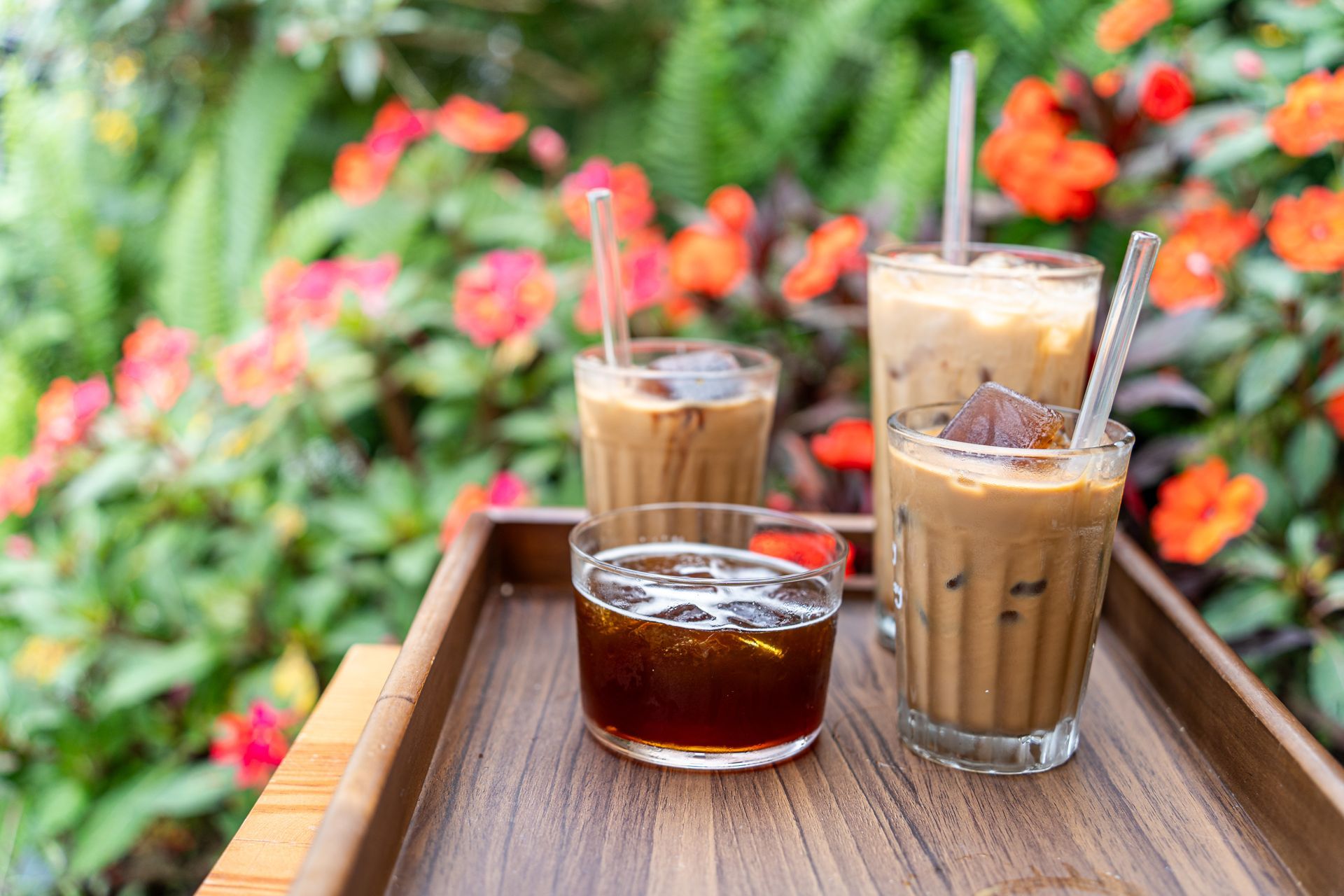 Four glasses of iced coffee, including one black coffee, on a wooden tray set against a background of bright orange flowers.