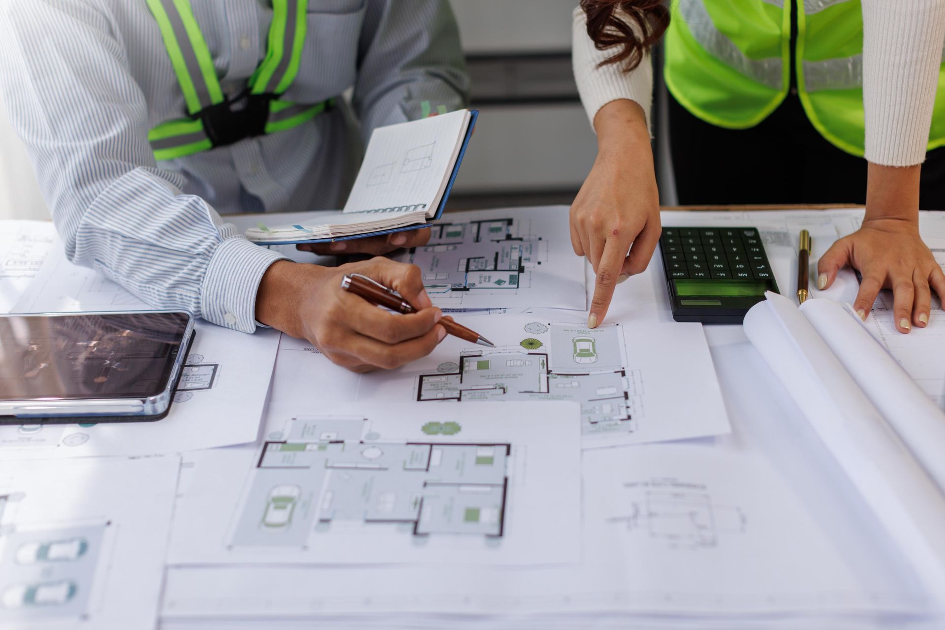 Two people wearing high-visibility vests review architectural floor plans on a table with a tablet, calculator, and pen.