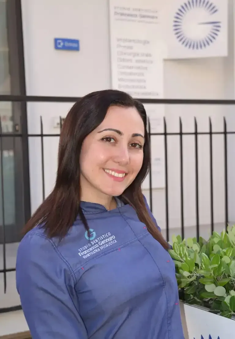 Donna sorridente con una camicia blu in piedi all'aperto accanto a delle piante e una recinzione nera.