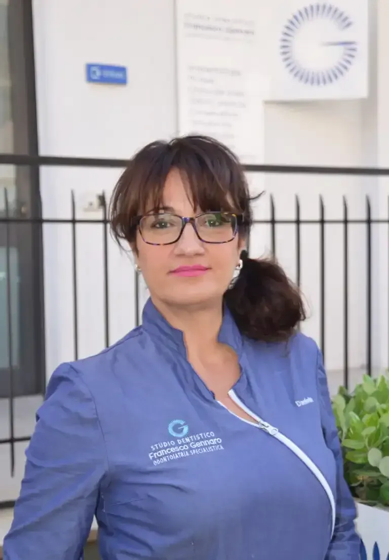 Una donna in uniforme blu è in piedi all'aperto davanti a un edificio, indossa gli occhiali e ha un leggero sorriso.