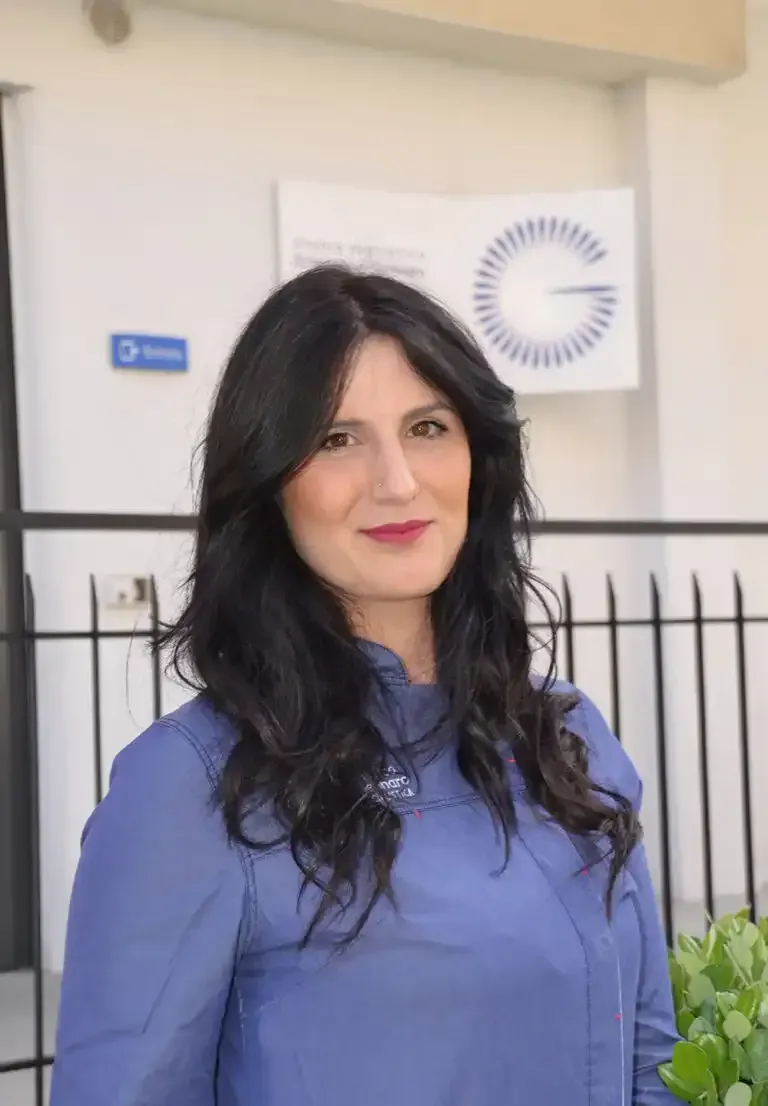 Una donna con una camicetta blu è in piedi all'aperto davanti a un edificio e sorride alla macchina fotografica.