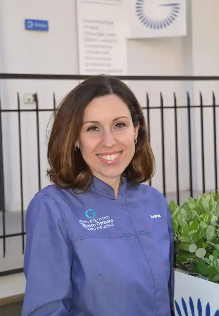 Persona sorridente in uniforme viola in piedi in un ambiente interno, vicino a una pianta e a una ringhiera nera.