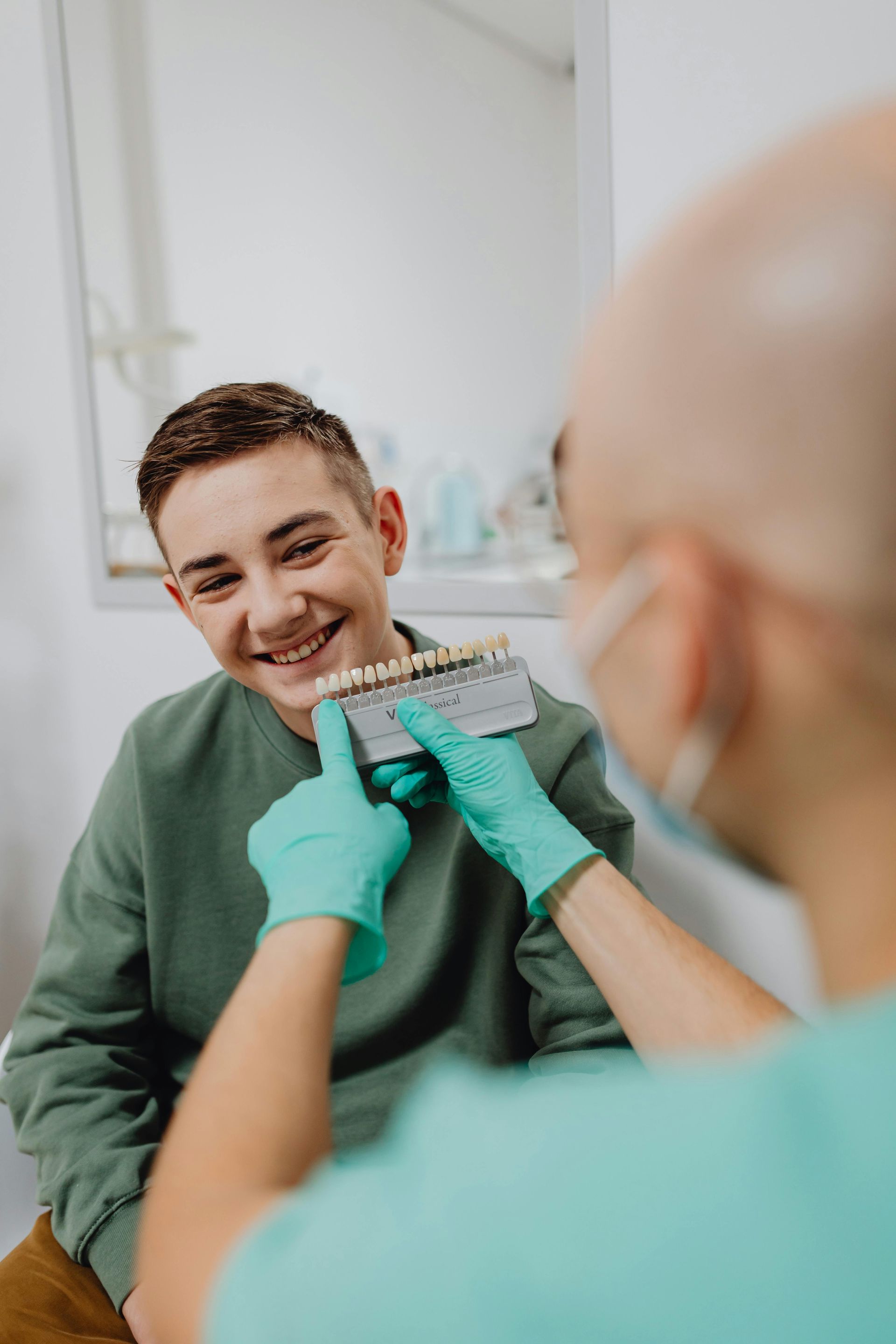 Un dentista tiene in mano un modello di denti mentre un paziente sorridente è seduto in una clinica odontoiatrica; si vedono i guanti verdi.