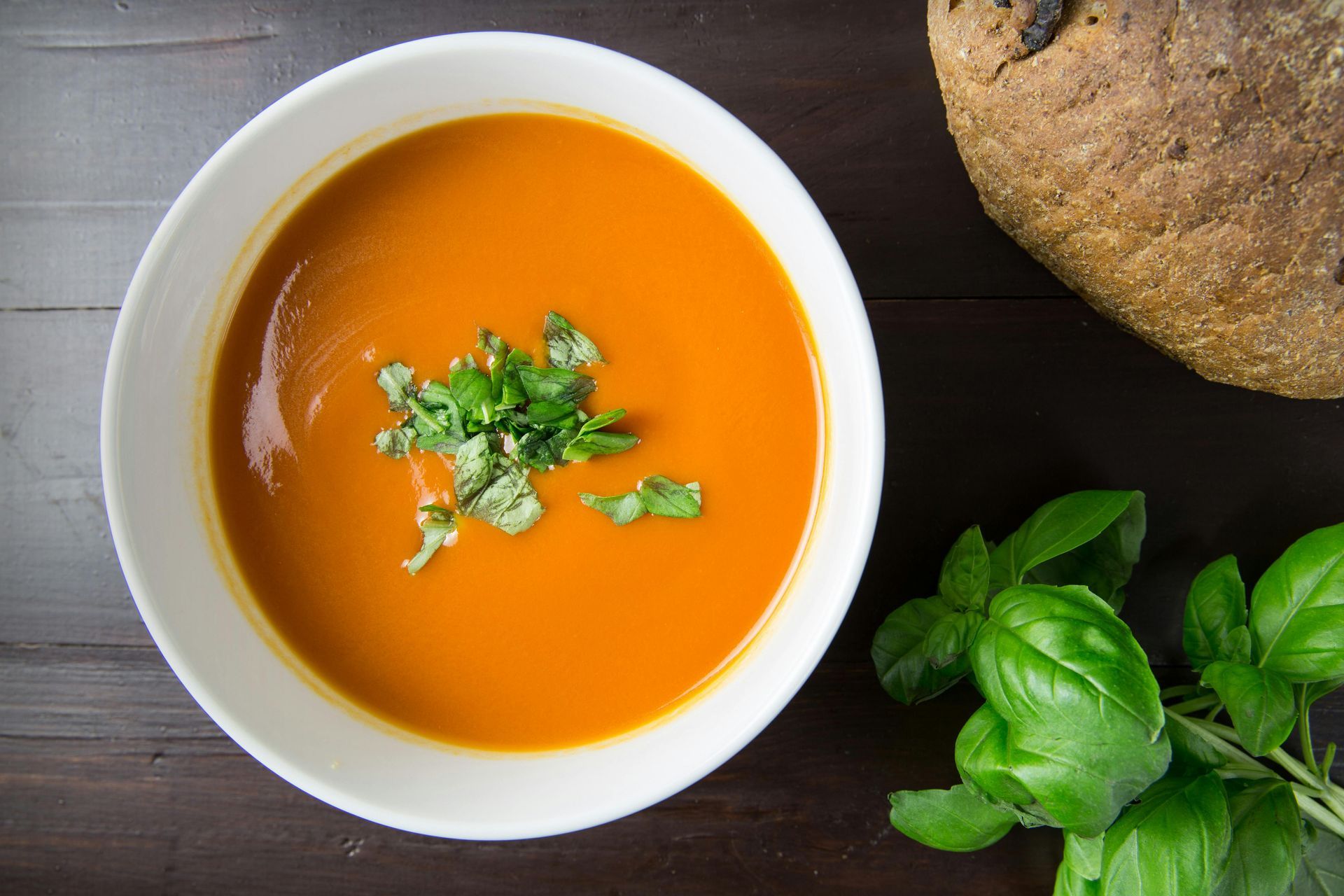 A bowl of tomato soup is sitting on a wooden table next to a loaf of bread.