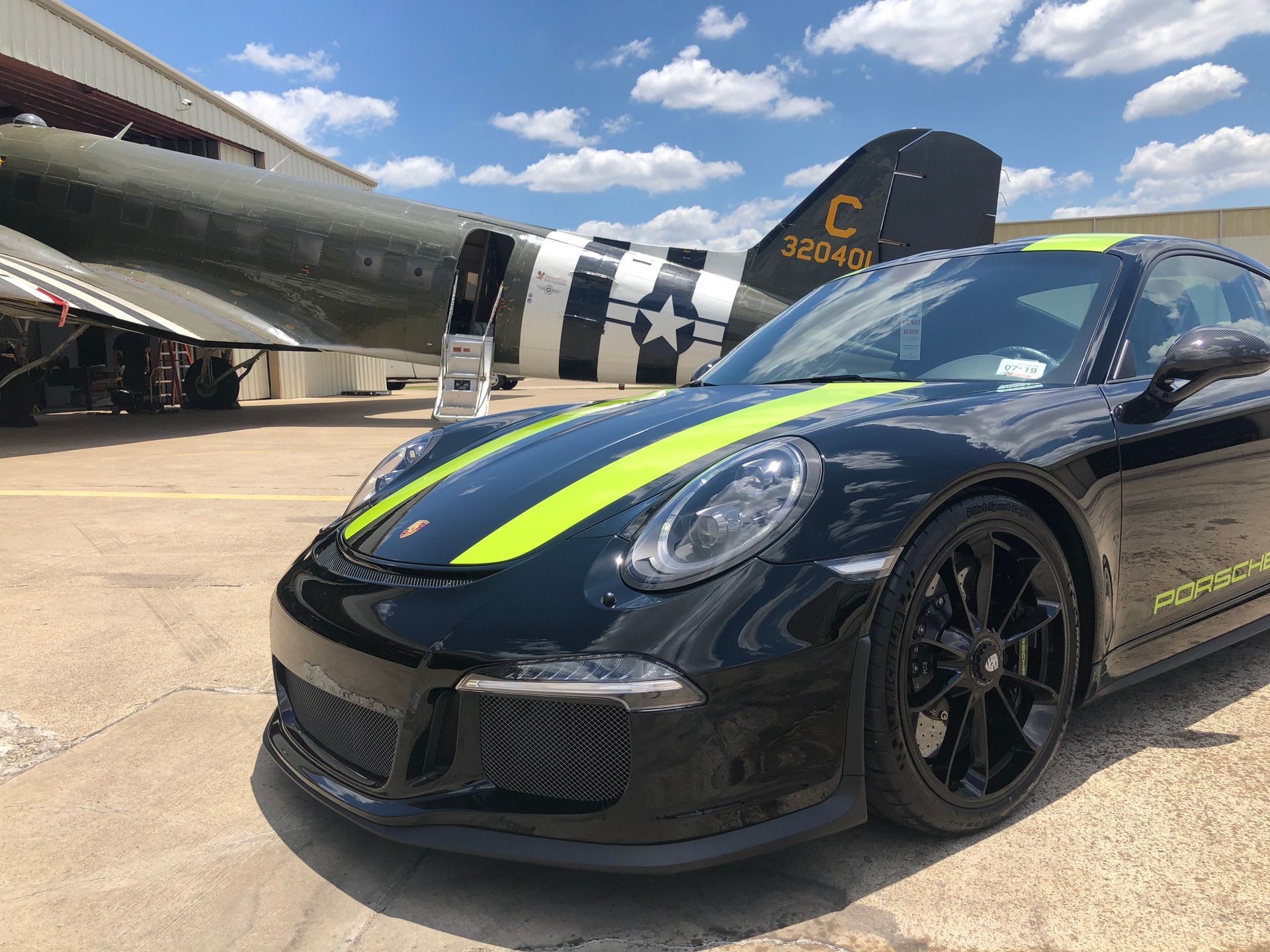 Black Porsche with neon green stripes parked in front of a WWII-era airplane.