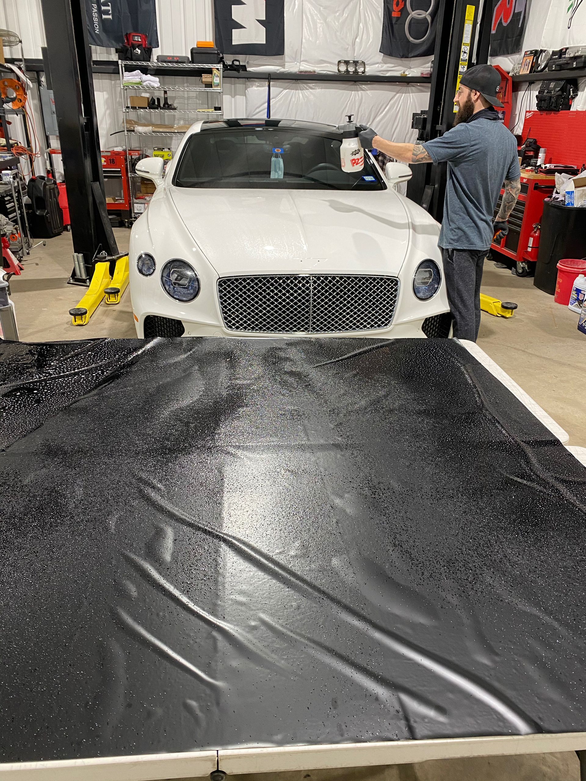 A person sprays a white Bentley in a garage; a black surface with glitter is in the foreground.