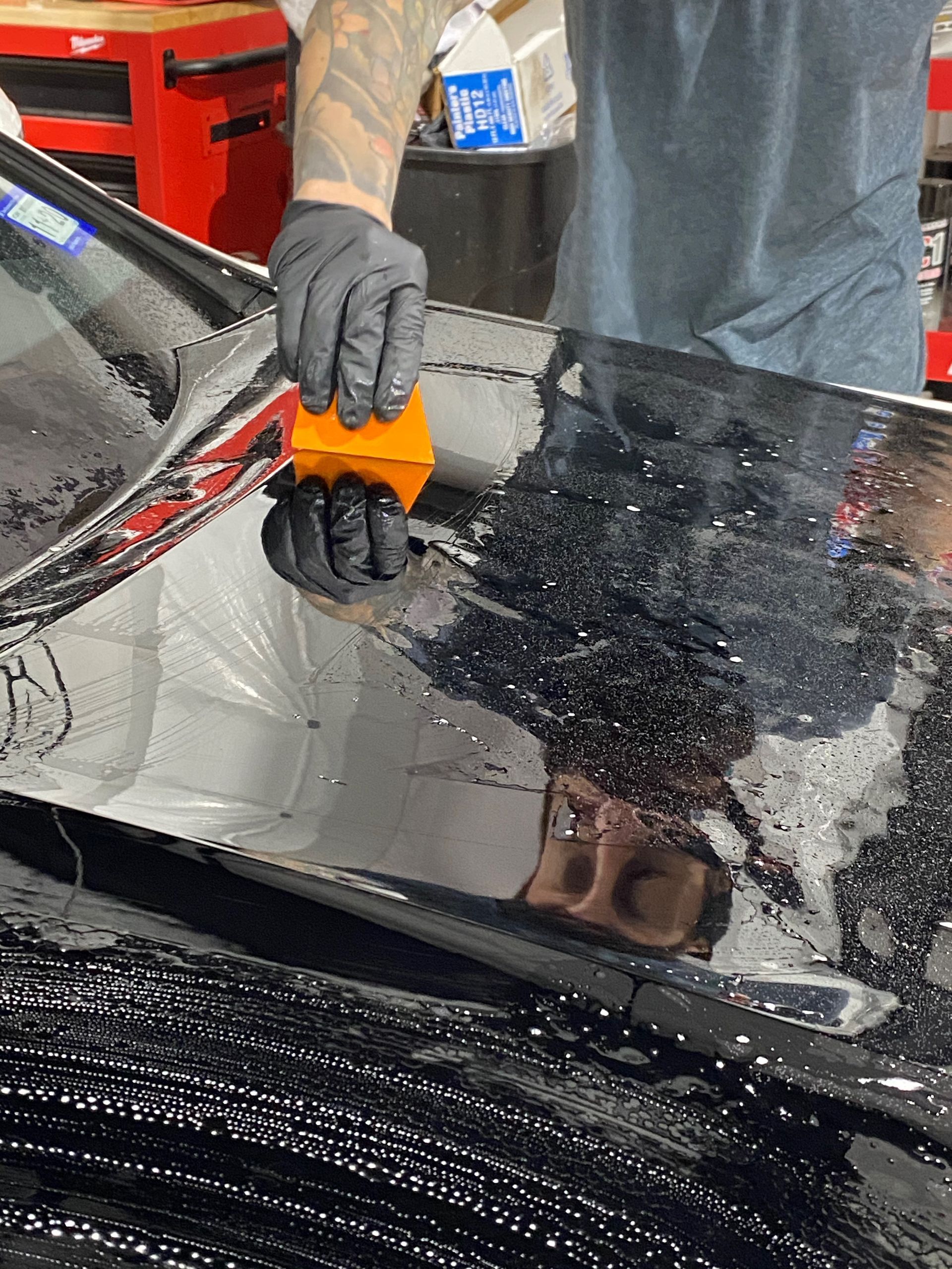 Person in black gloves applying window tint to a car window with an orange squeegee in a workshop.