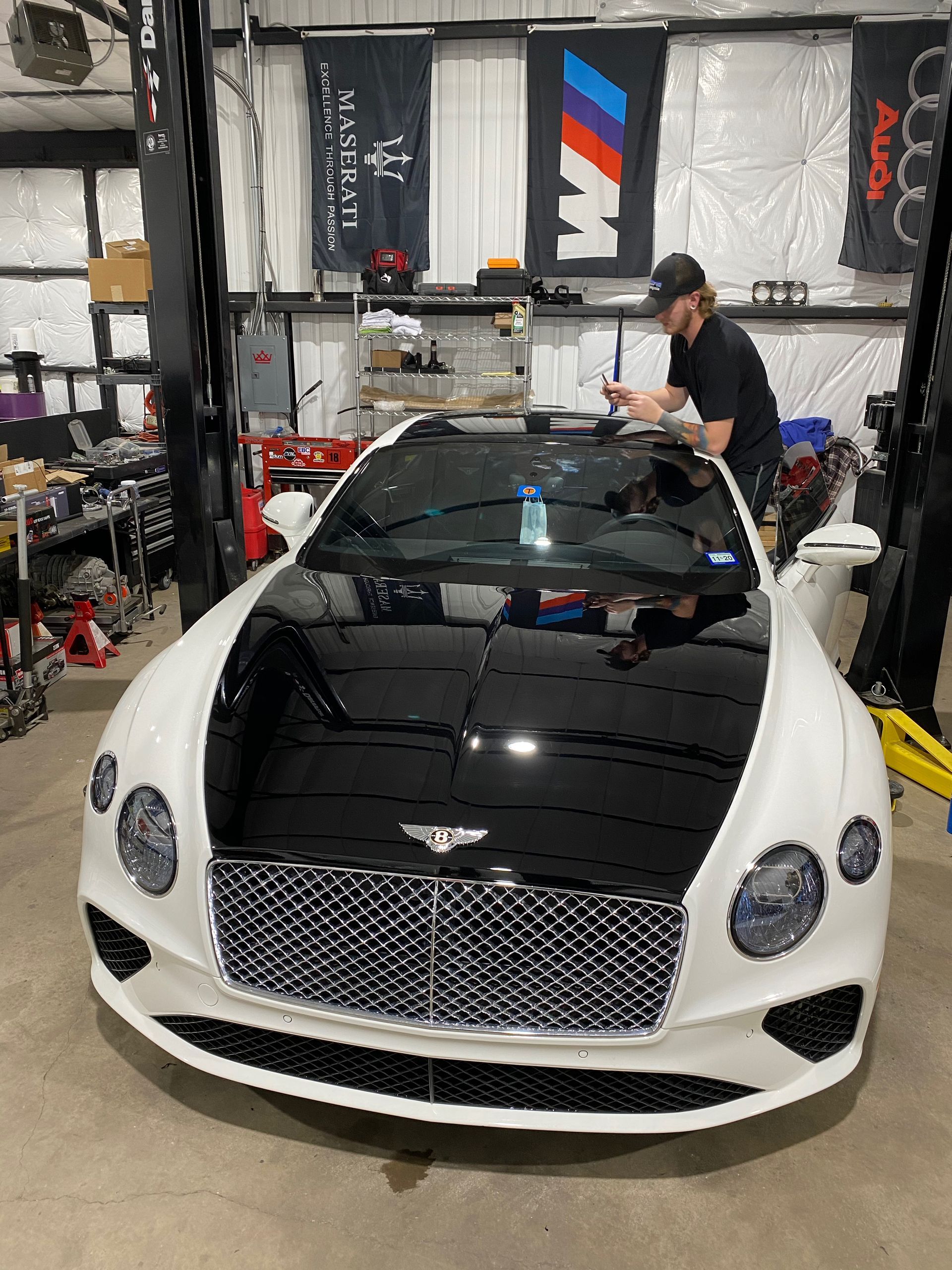 A white and black Bentley in a garage, person working on roof, flags in background.