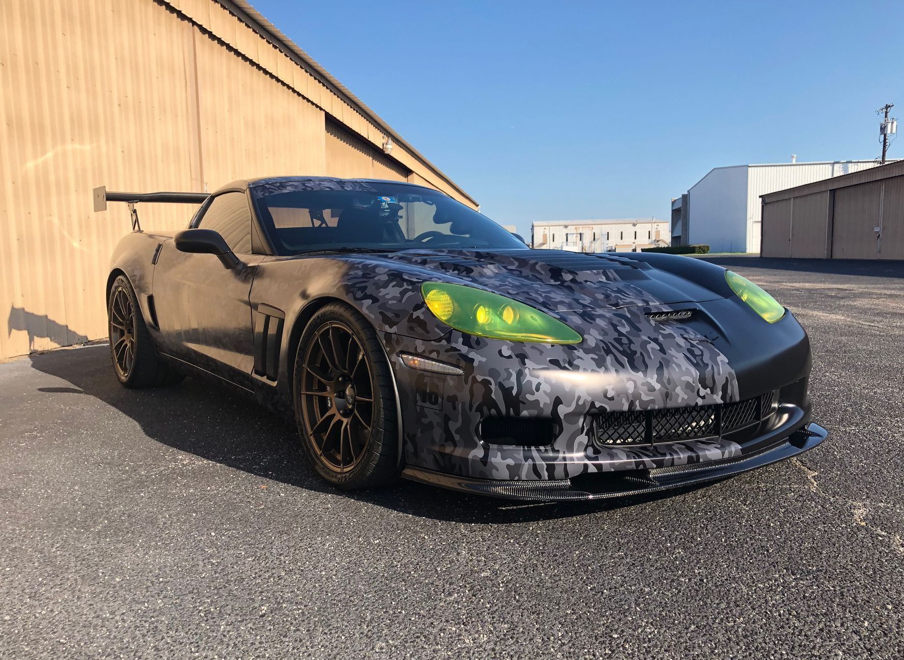 Black Corvette sports car with gray camouflage front, gold wheels, and large rear wing, parked outside.