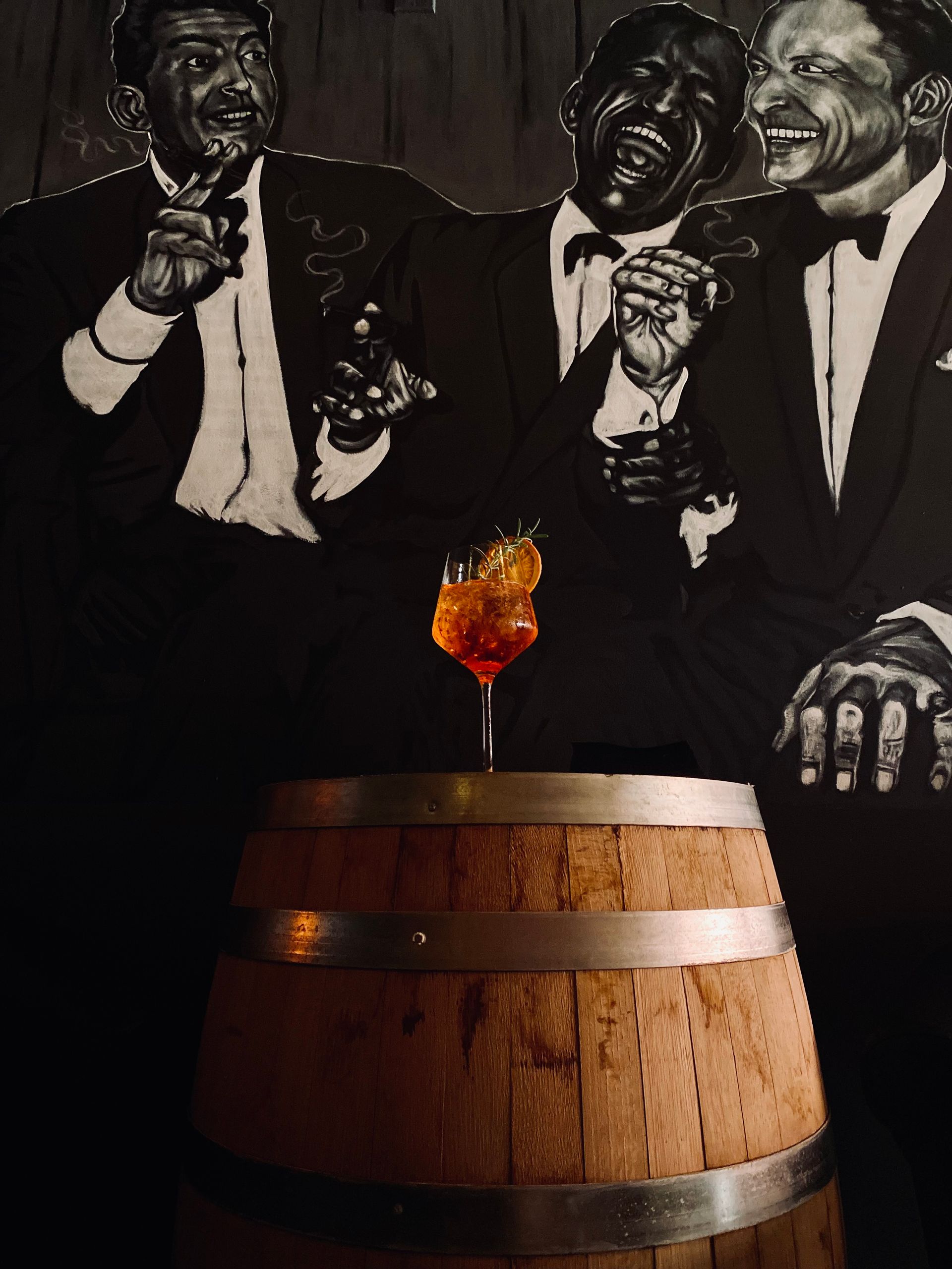 A barrel with a glass of wine on top of it, in front of a mural of the 'Rat Pack'