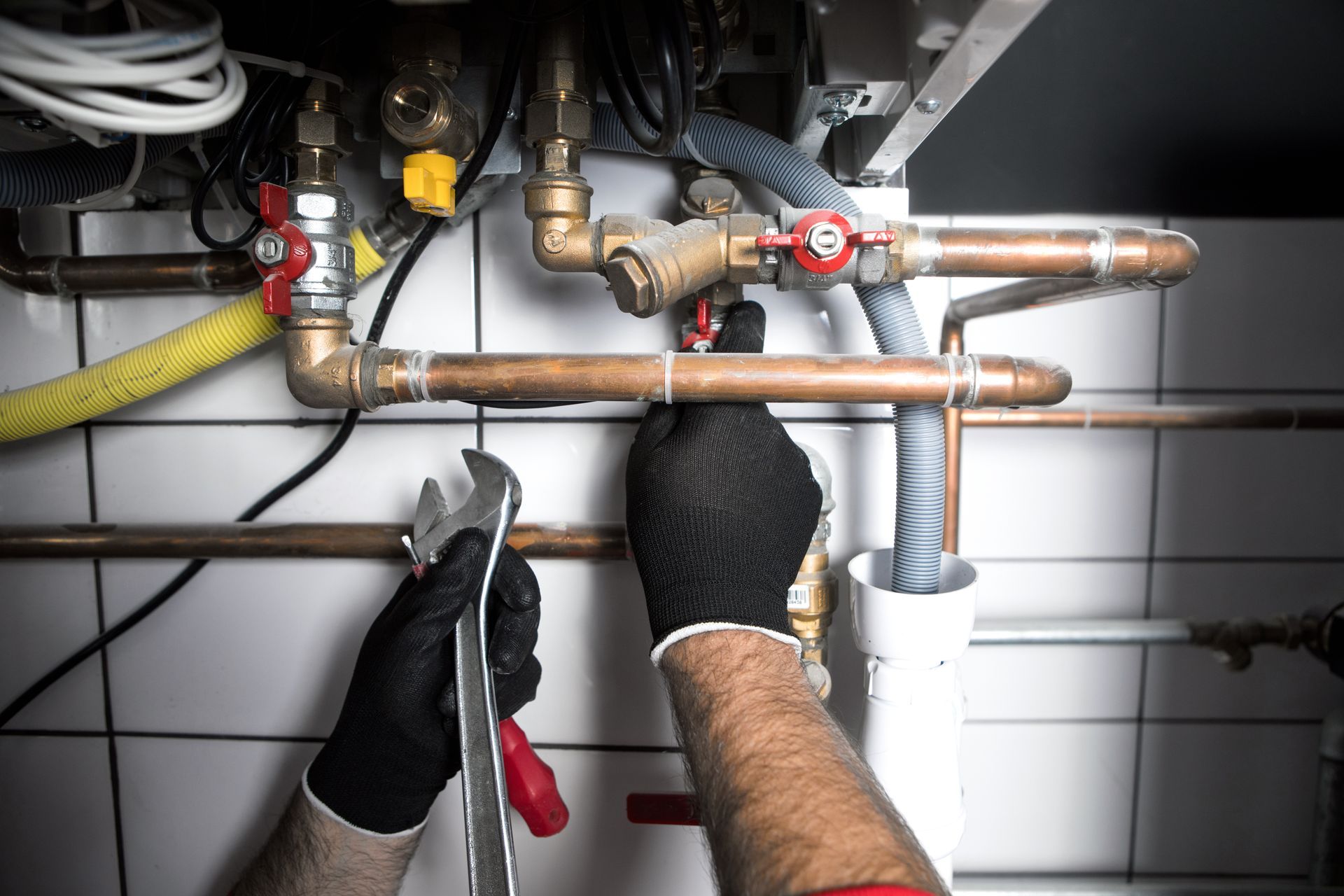 Gloved hands repairing copper pipes and valves under a sink with tools in a tiled utility area
