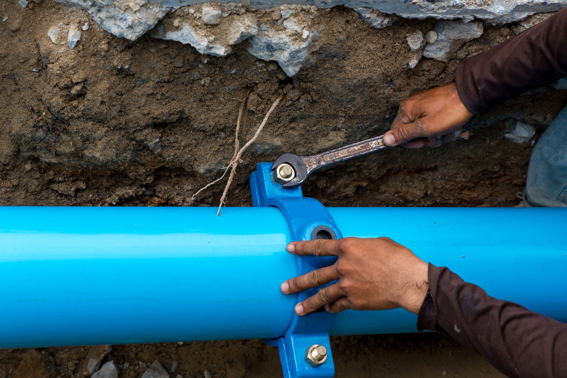 Hands tightening a blue pipe joint with a wrench against a dirt wall.