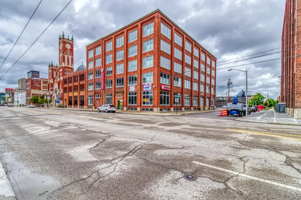 Brick building on a city street with a tower in the background under a cloudy sky.