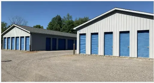 Two gray storage buildings with blue doors stand on a gravel lot under a bright blue sky.