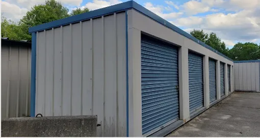 Row of storage units with blue doors, a concrete floor, and a blue-trimmed roof. Trees are visible in the background.