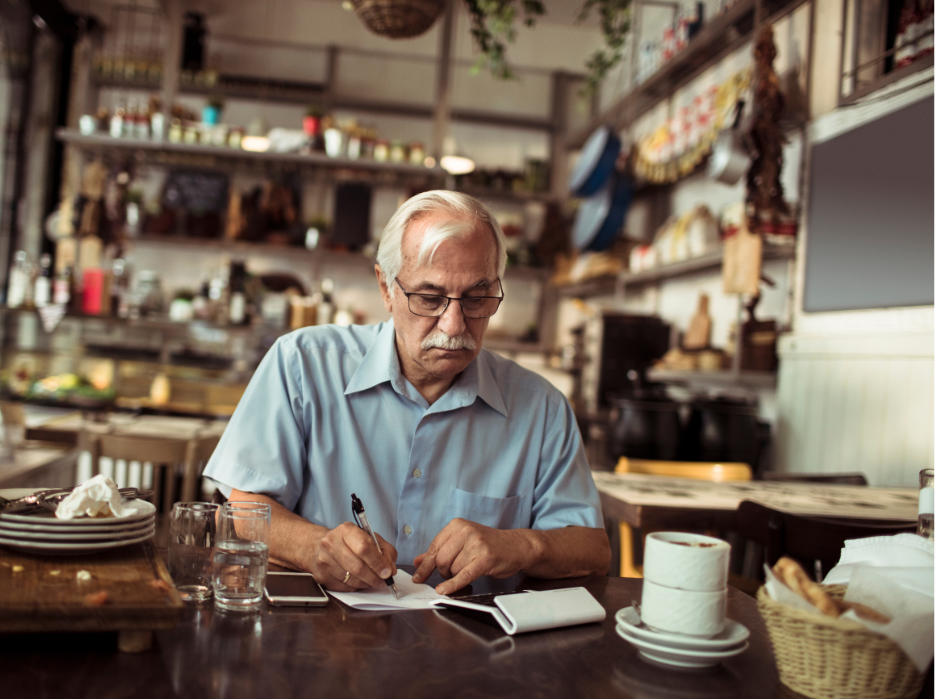 An older man is sitting at a table in a restaurant writing in a notebook.