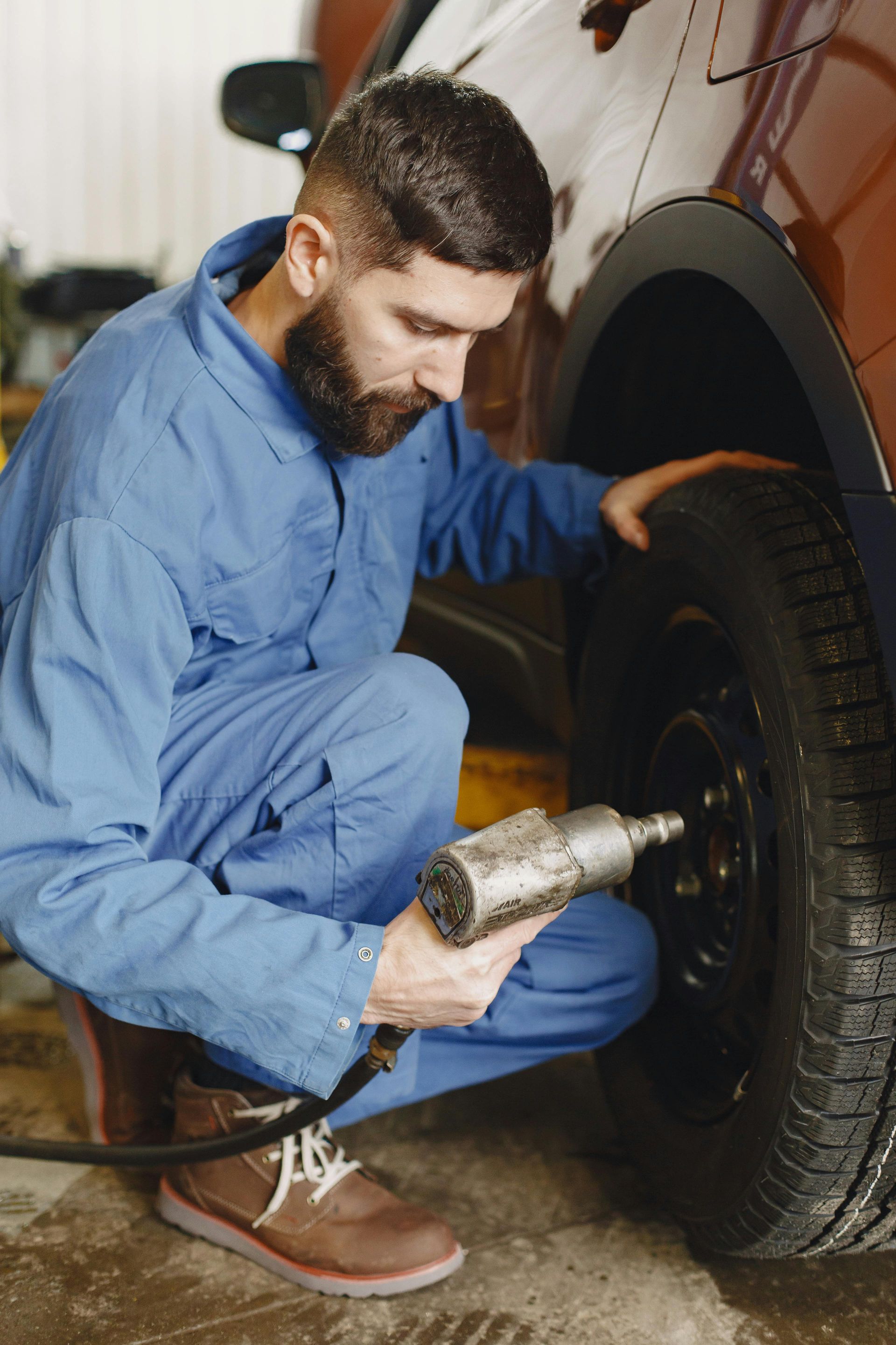 Mechanic in blue coveralls, uses a power tool to work on a car tire at a garage.