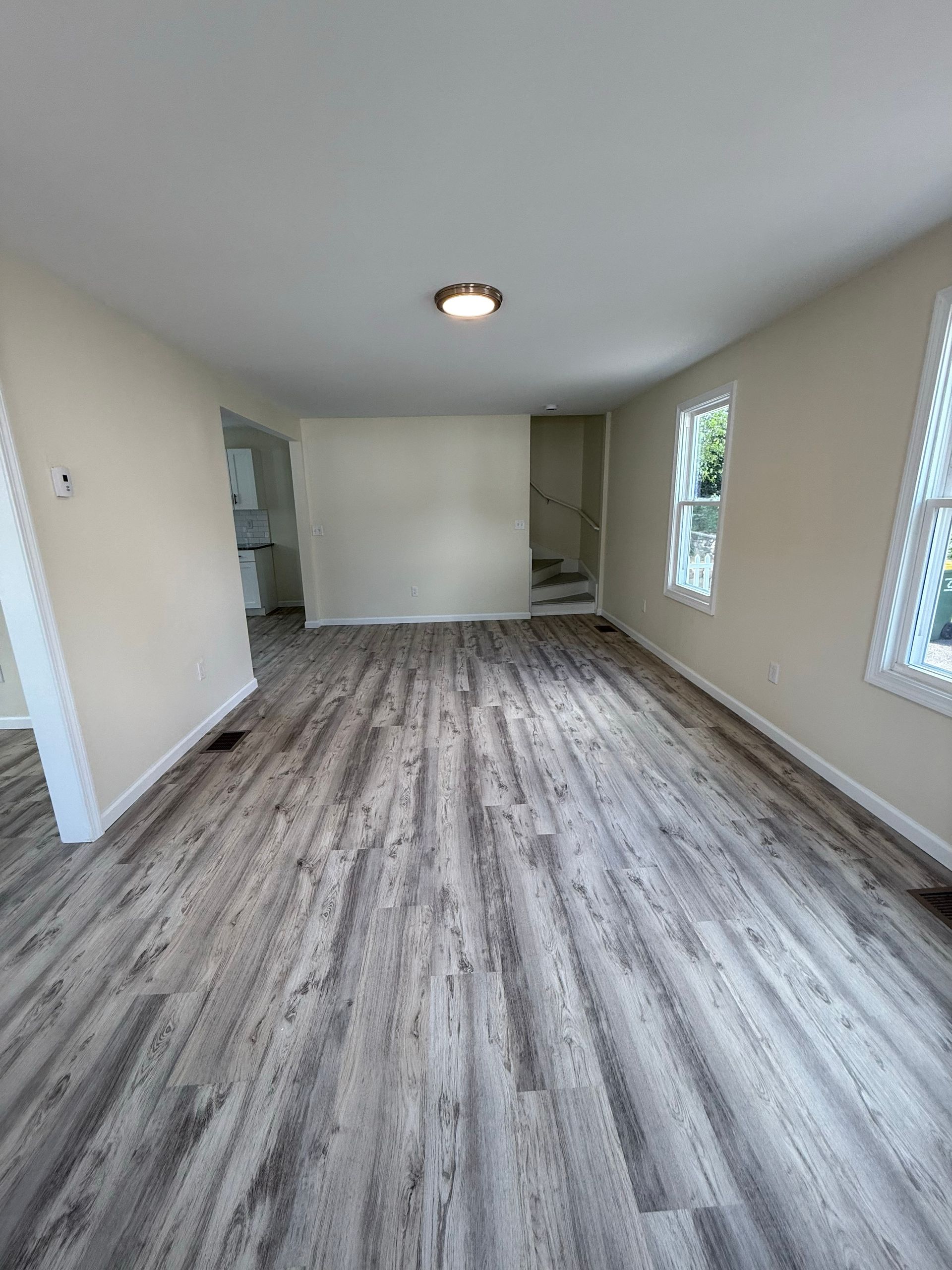 Empty room with gray and white wood-look flooring, beige walls, and white trim. A single light fixture is centered in the ceiling.