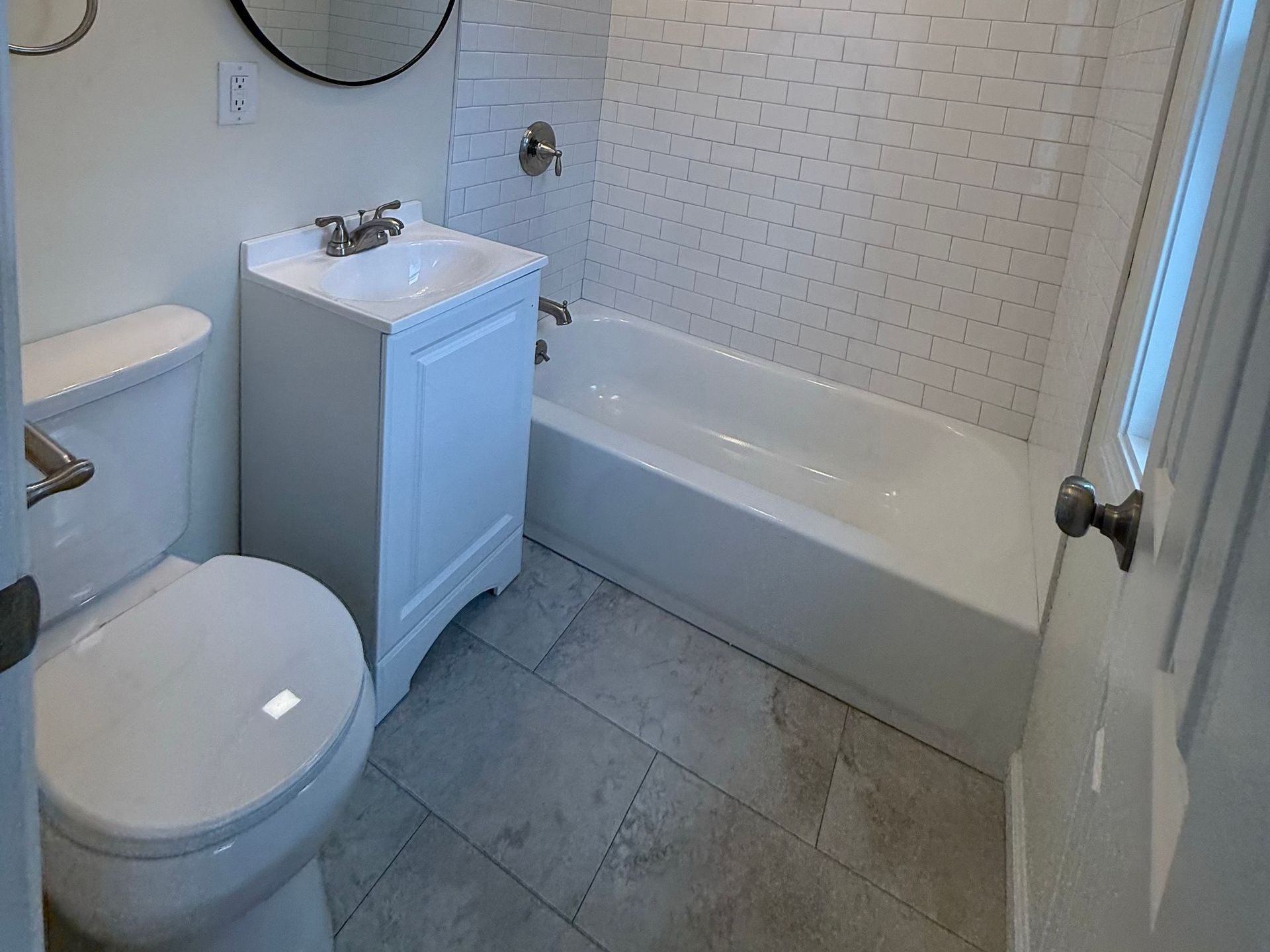 A small, recently renovated white bathroom with a toilet, vanity, and bathtub; gray tiled floor and white wall tiles.