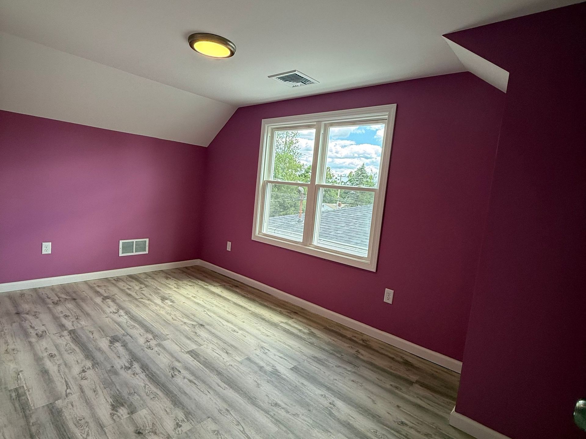 A bedroom with purple walls, a window, and light gray wooden flooring. The room has a sloped ceiling.
