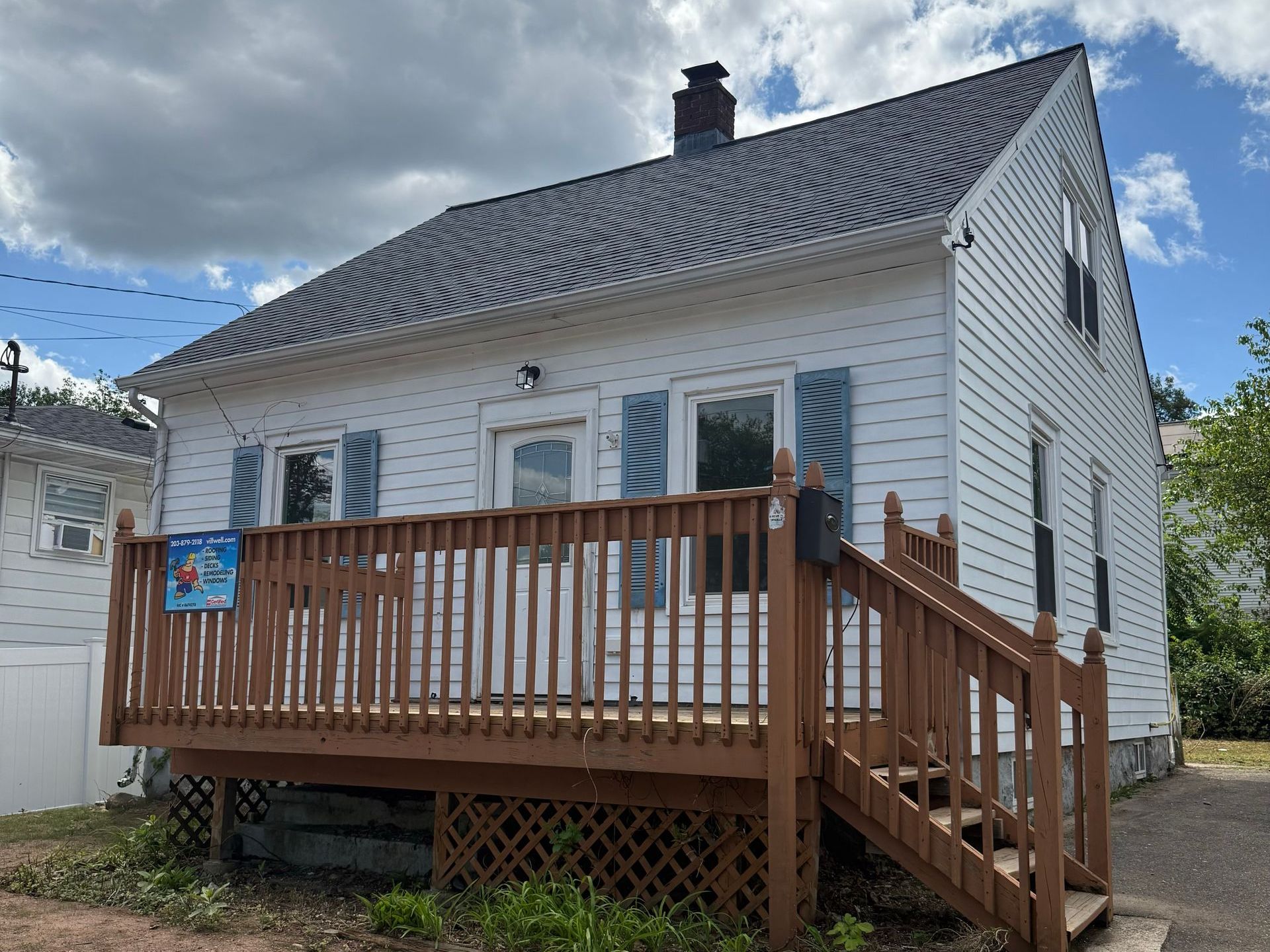White house with a brown wooden deck, blue shutters, and a dark roof under a partly cloudy sky.