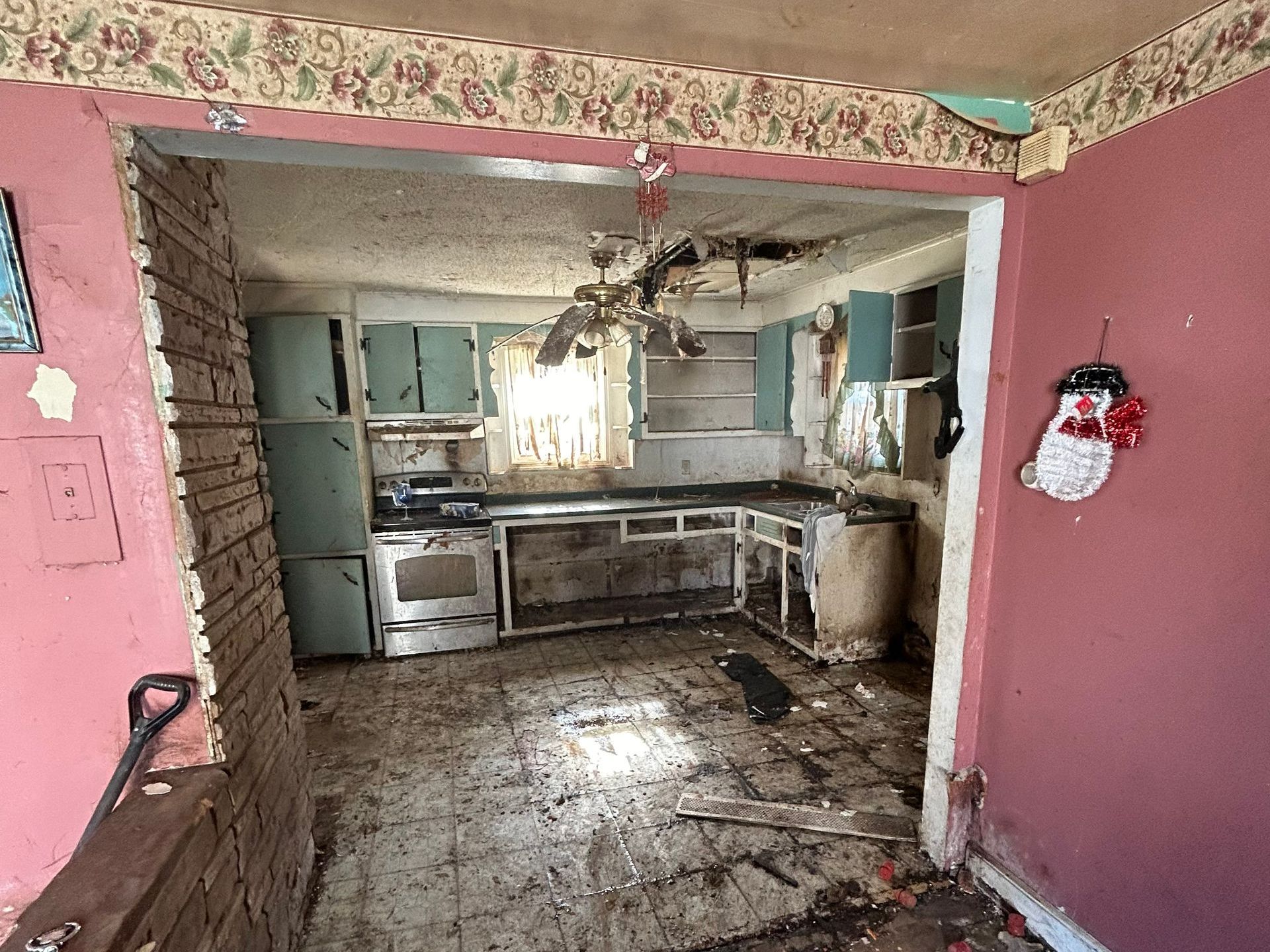A dilapidated kitchen with light blue cabinets, stained appliances, and debris on the floor. A doorway leads to the kitchen.